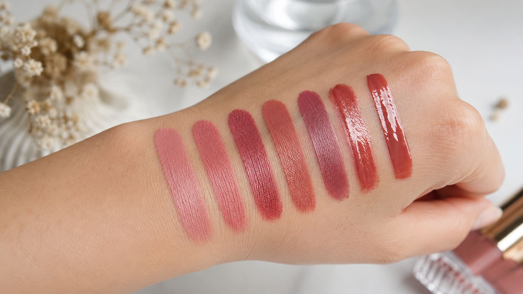 A hand with seven different swatches of various nude and pink lipstick shades on the skin.