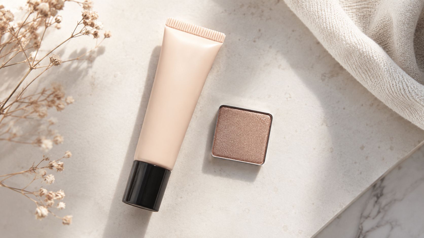 A beige tube of primer and a small square pan of shimmering eyeshadow on a neutral surface.