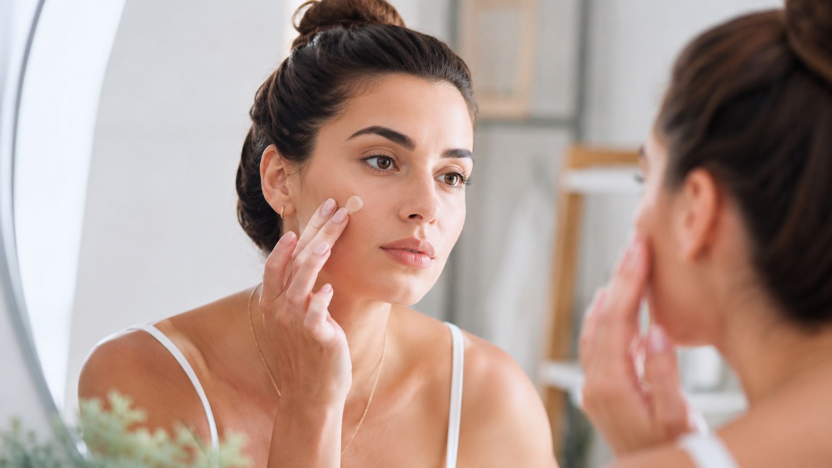 A woman applying a small dot of skincare product or concealer to her cheek while looking in mirror.