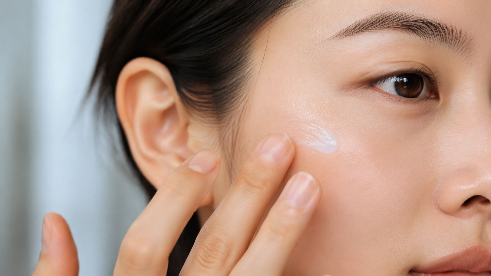 A close-up of a person applying a smooth skincare cream to their cheek with their fingertips.