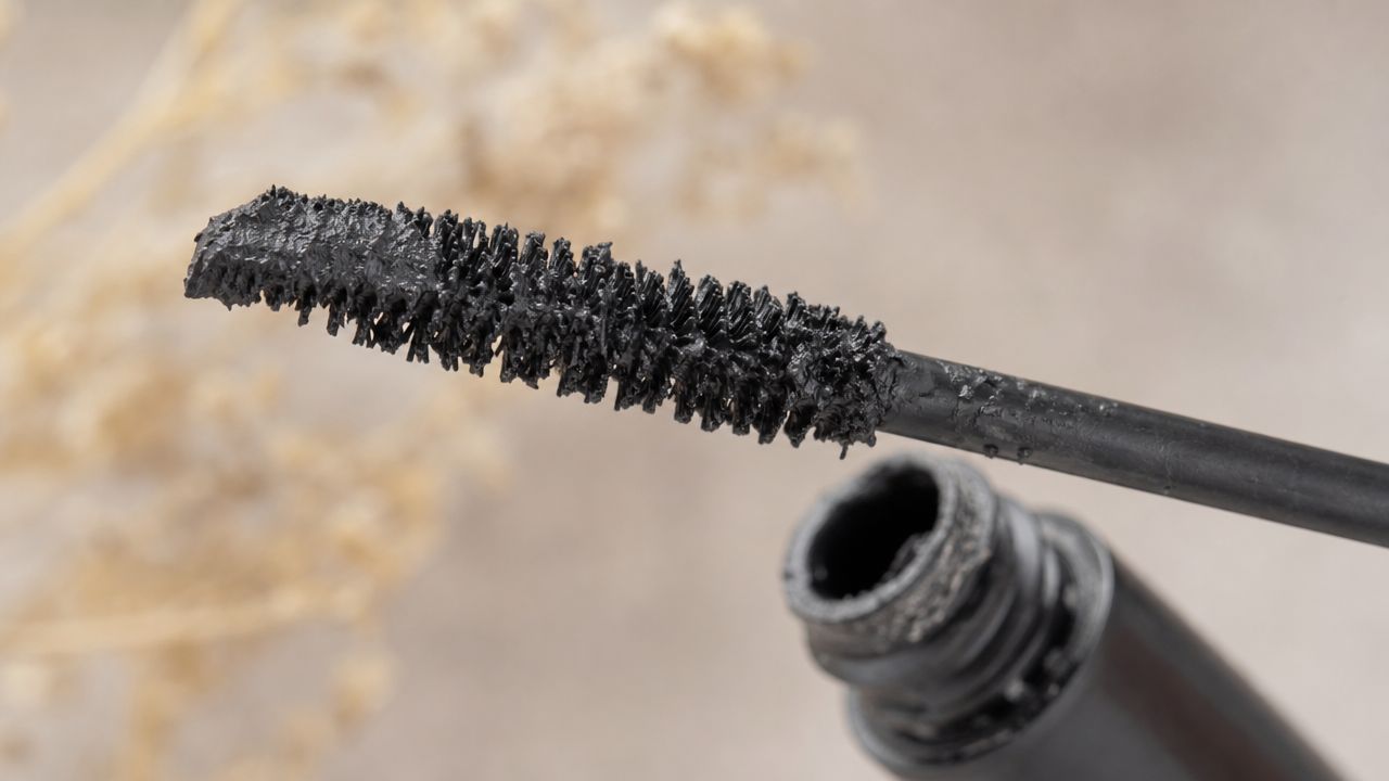 A close-up view of a black mascara wand coated in thick product next to its open tube.