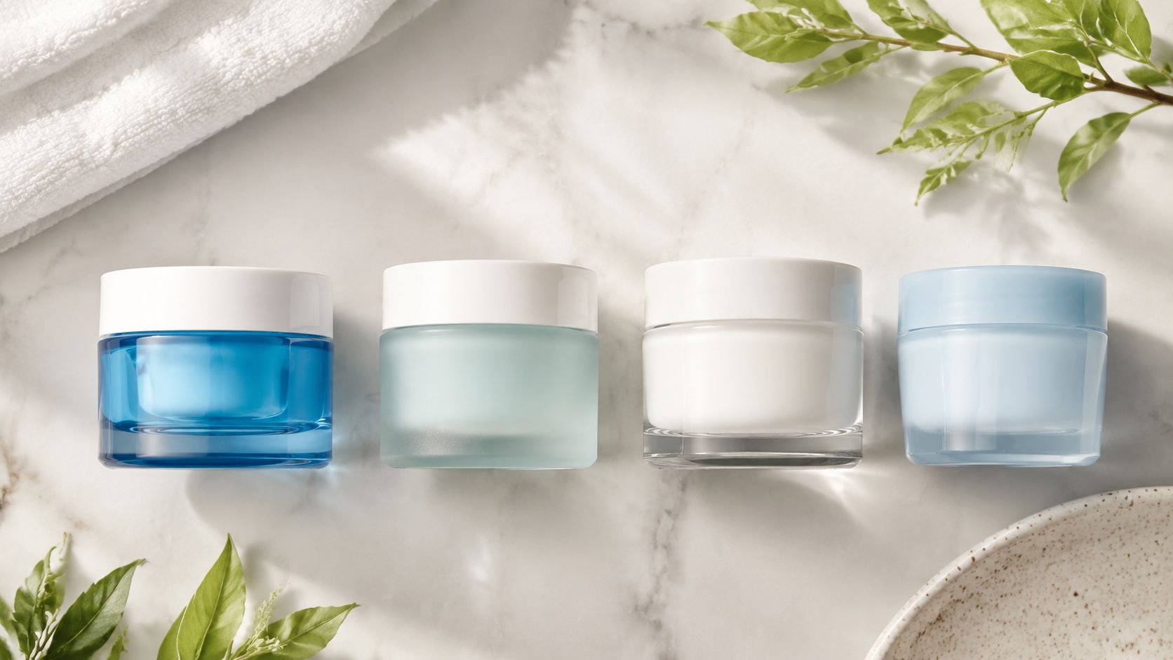 Four different cosmetic cream jars arranged on a marble surface next to a white towel and leaves.