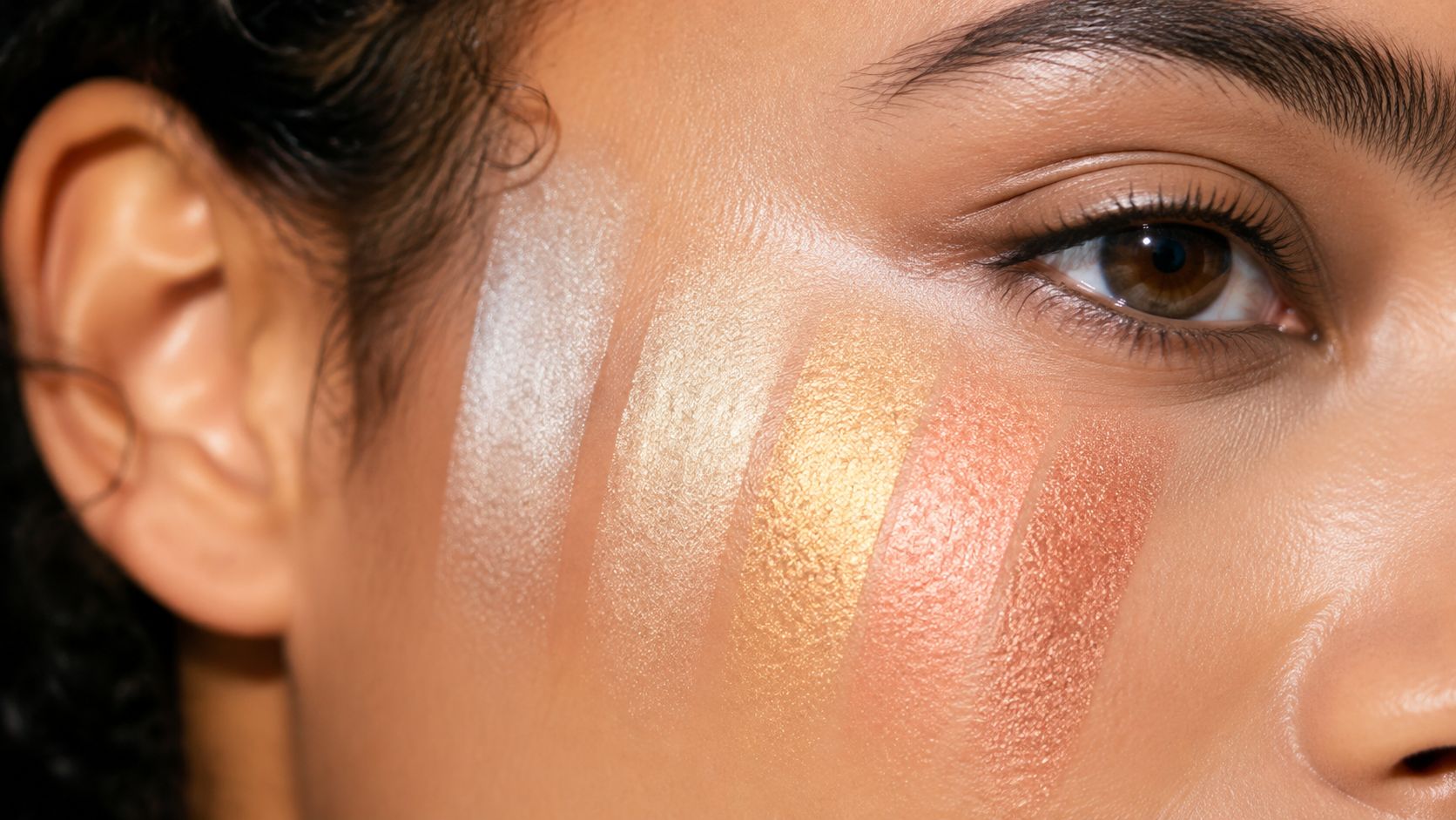 Close-up of a person with five shades of shimmering highlighter swatched on their cheekbone.