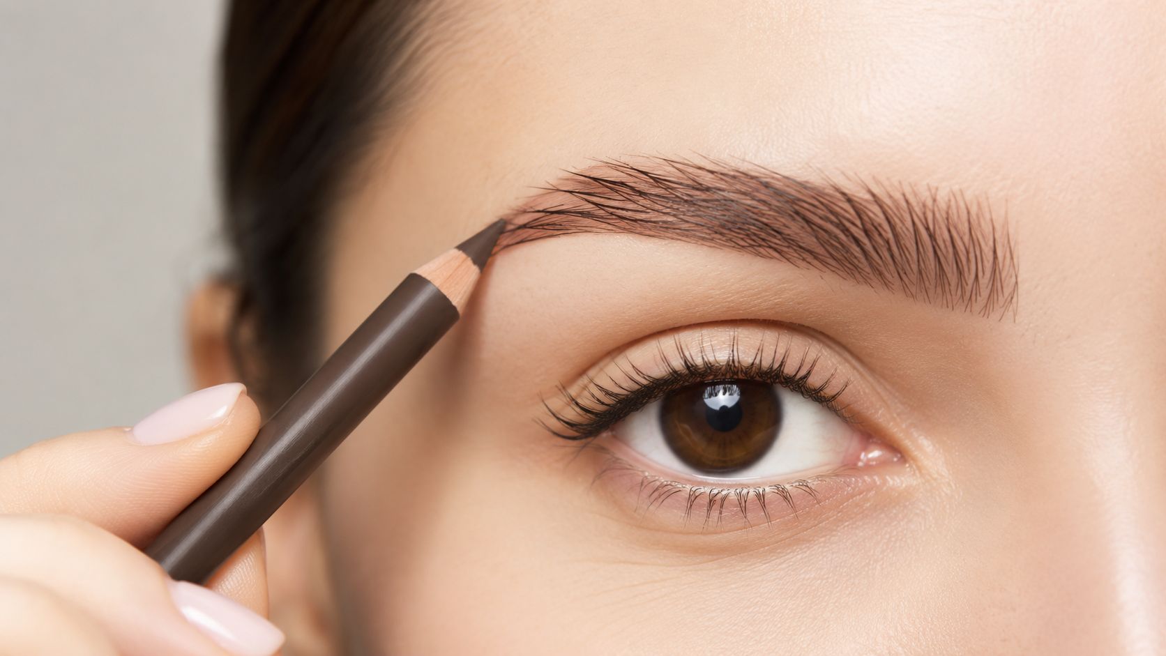 A close-up view of a woman using a dark brown cosmetic pencil to shape her eyebrow.