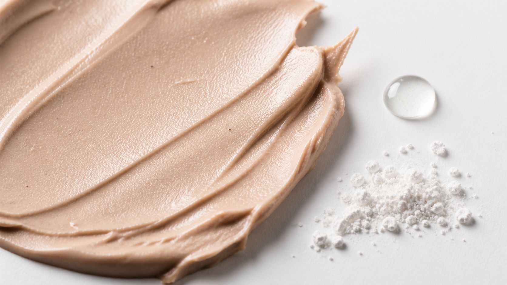 A beige cosmetic cream swatch next to a clear droplet and white cosmetic powder on white background.