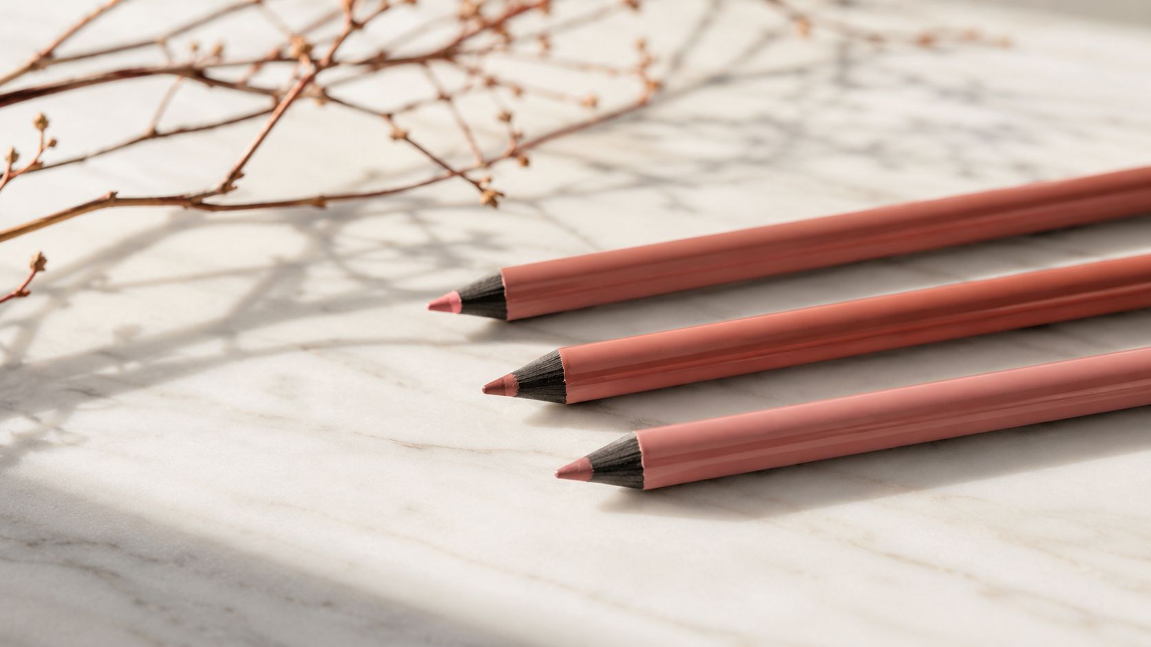 Three peach-toned cosmetic lip pencils arranged neatly on a marble surface with decorative twigs in the background.