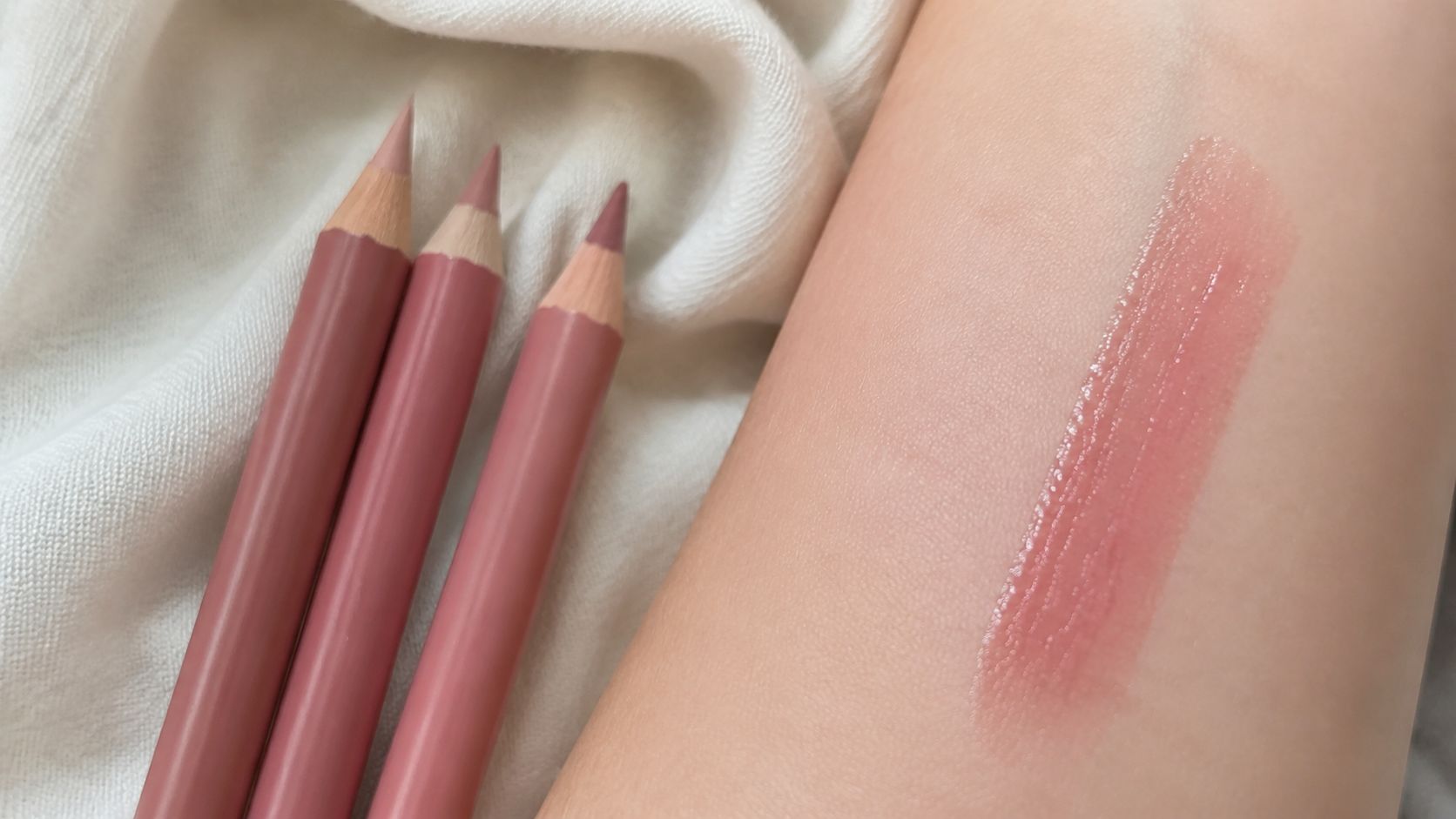 Three dusty rose lip pencils lined up next to a glossy pink swatch on skin