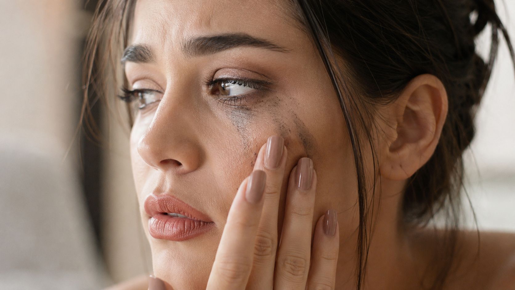 A distressed woman with smudged mascara and makeup running down her cheek touching her face.