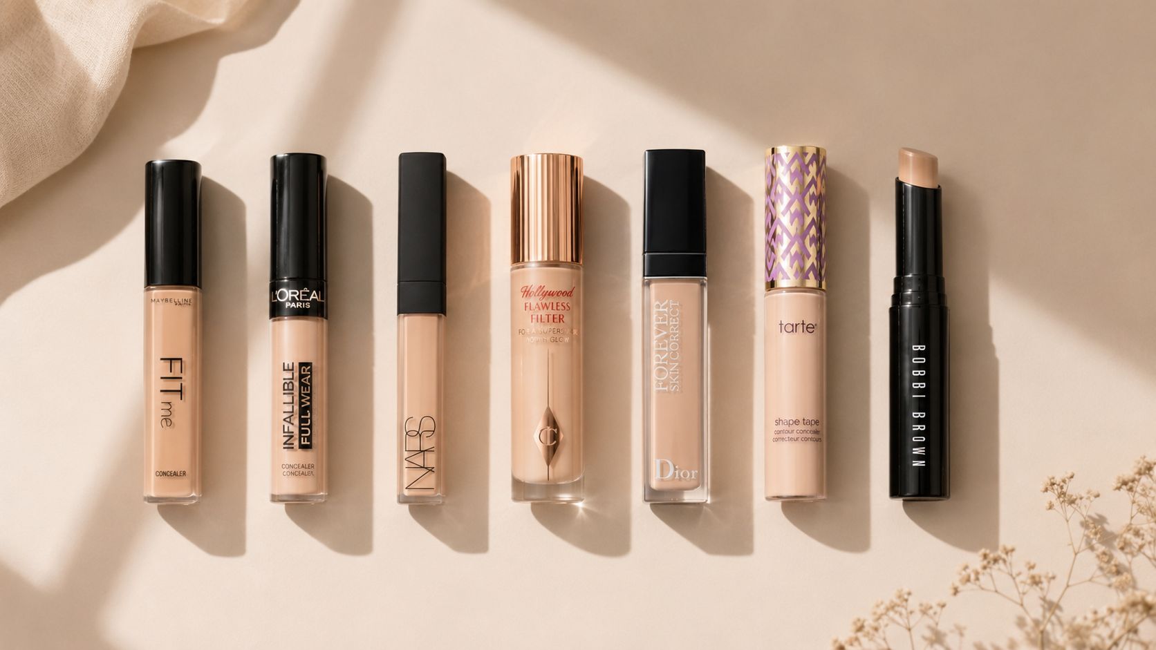 A line of various high-end and drugstore concealer bottles arranged neatly on a light beige background.
