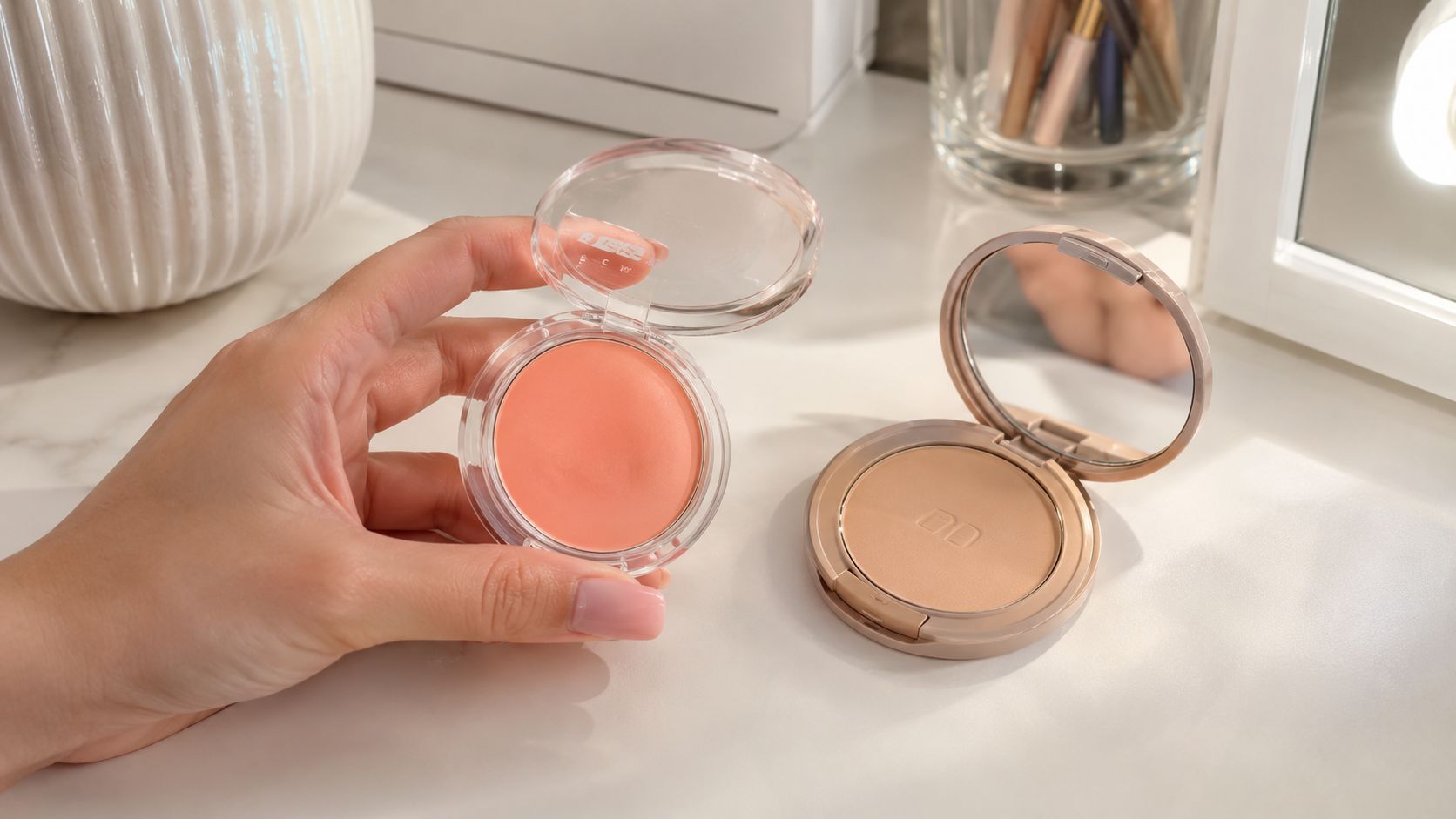 A hand holds an open container of peach cream blush next to a compact powder mirror.
