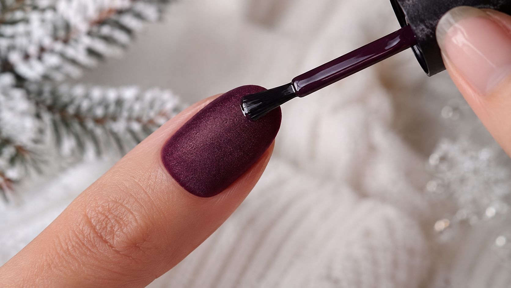 A close-up of a person applying dark burgundy matte nail polish onto their fingernail during winter.
