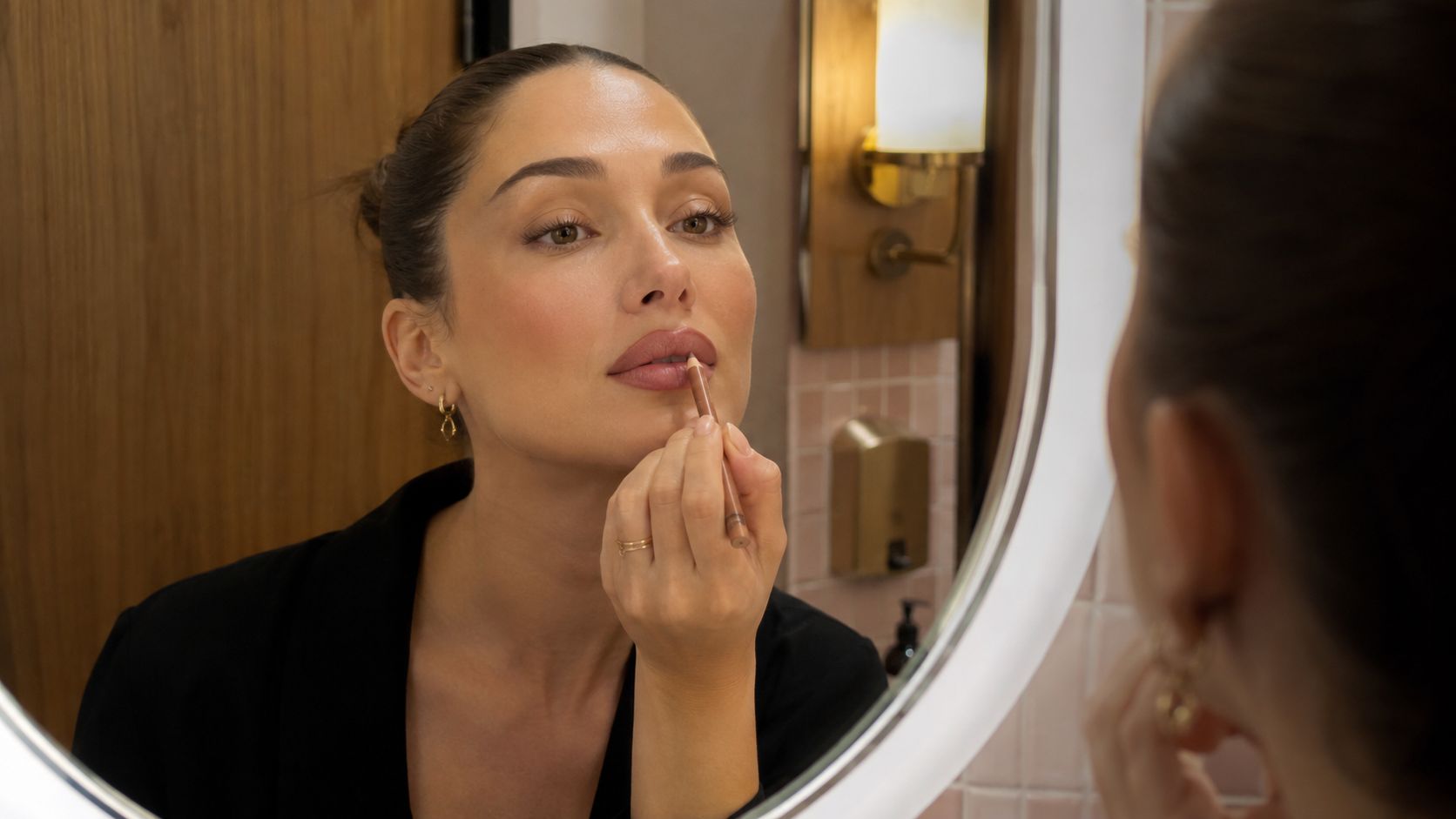 A woman looks in the bathroom mirror while carefully applying lip liner to her lips.