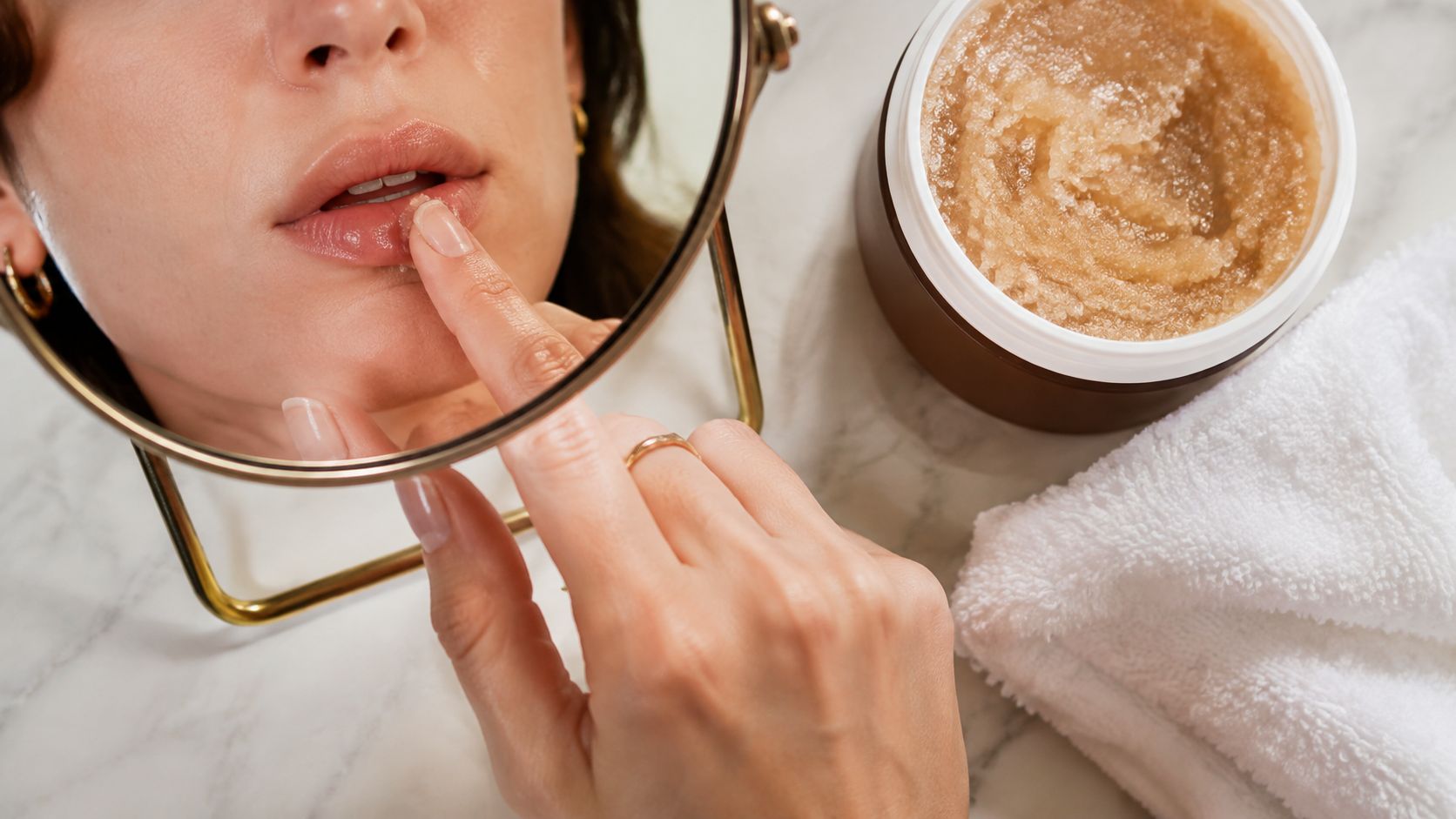 A woman applying a sugar lip scrub from a jar onto her lips while looking in a mirror.