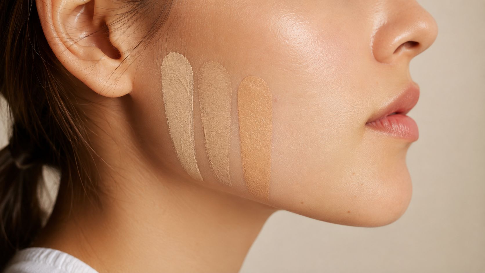 A close-up view of a woman with three different shades of concealer swatched on her jawline.