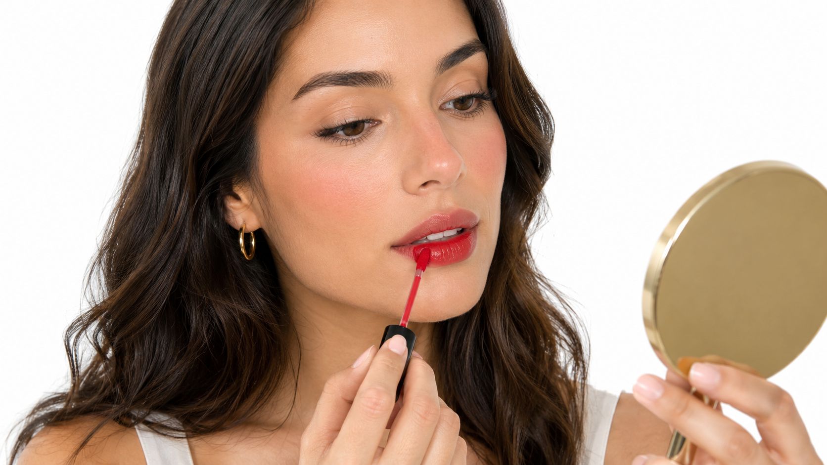 A woman applying vibrant red lip stain to her lips while looking into a handheld gold mirror.