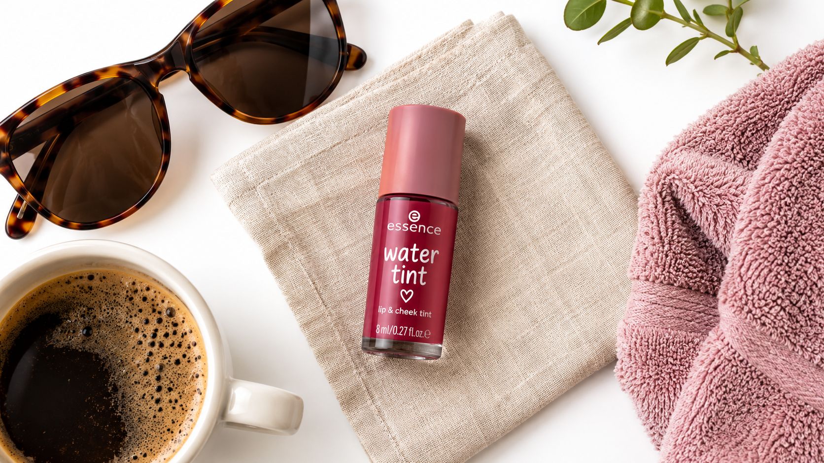 A small bottle of Essence water lip and cheek tint resting on a linen cloth next to coffee and sunglasses.