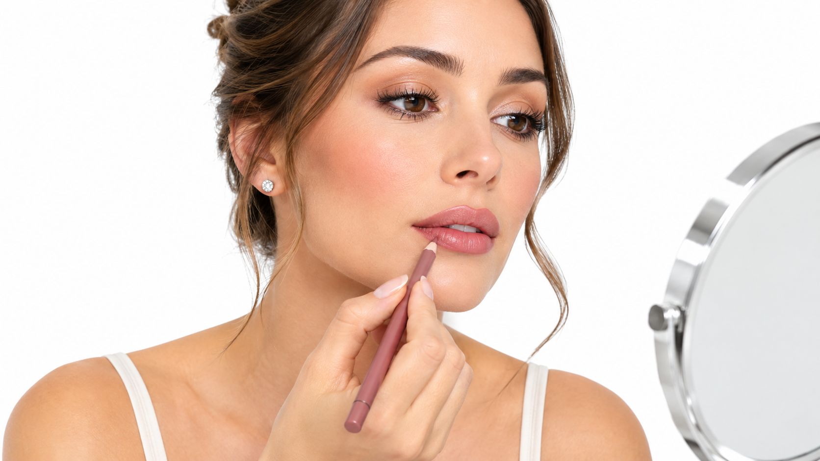 A woman applying a pinkish nude lip liner pencil to her lips while looking into a mirror.